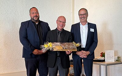 Herr Miller mit Herrn Knauer und Herrn Kiesel, er hält ein Geschenk in der Hand. 